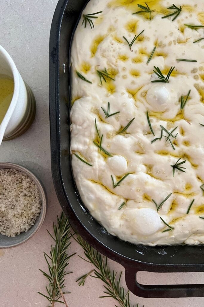 A sourdough focaccia loaf sitting in a cast iron bread pan. The dough is bubbly and covered with salt, rosemary and olive oil. It is unbaked.