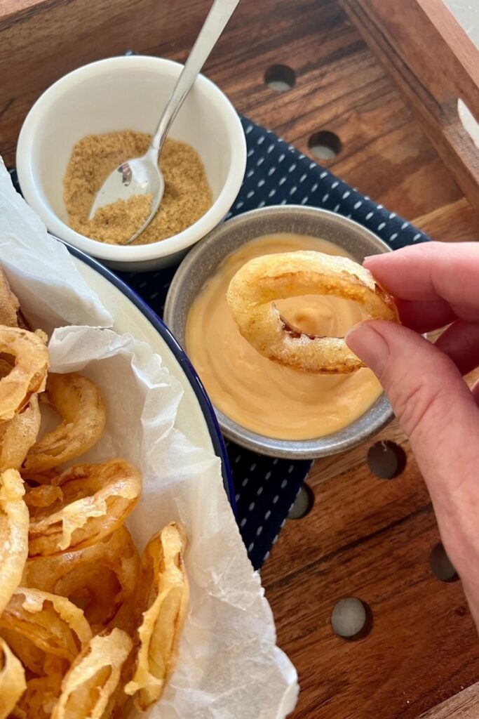 Sourdough onion ring being dipped into a dish of kewpie mayo blended with sriracha hot sauce. There's also a dish of seasoned salt sitting on a blue dish towel.