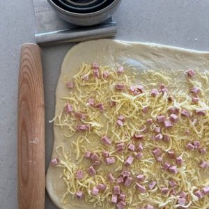 dough sprinkled with cheese and ham pieces. There is a dough scraper, bowl and rolling pin sitting next to the dough in the photo.