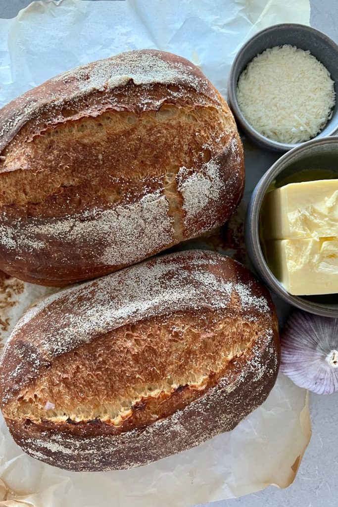 2 small sourdough loaves pictured with a head of garlic, bowl of butter and bowl of parmesan cheese.