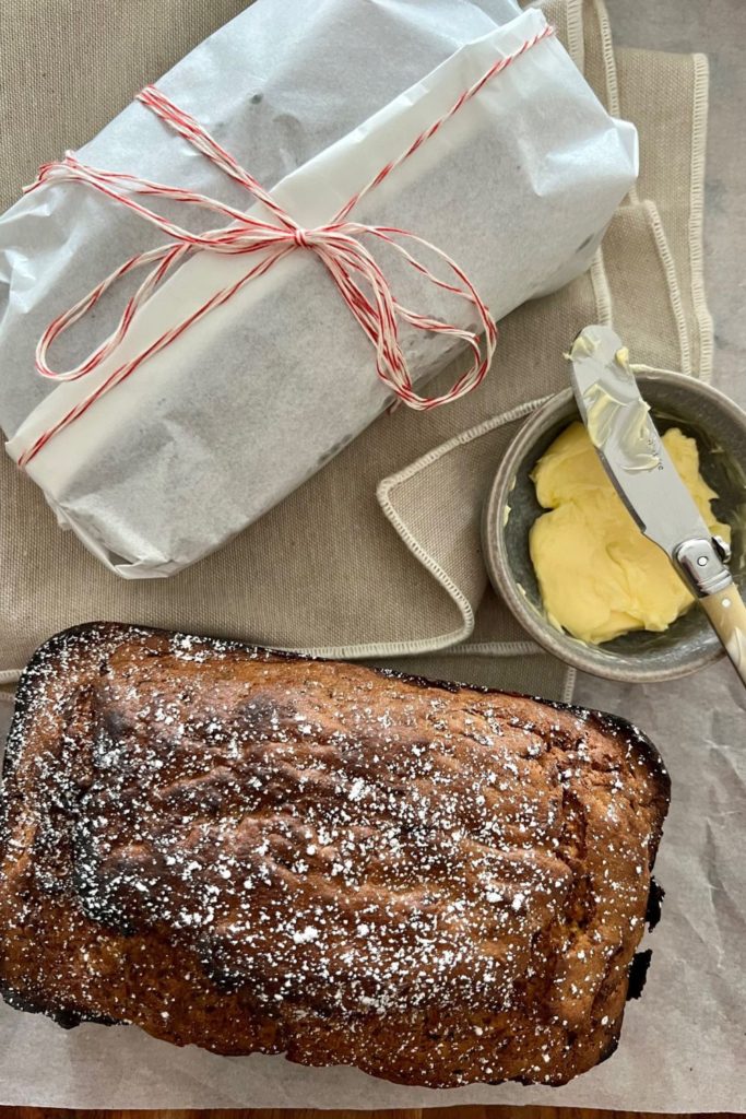 One loaf of sourdough zucchini bread wrapped in wax paper tied with red string. Another loaf is dusted in icing sugar. There is a bowl of butter sitting between them.