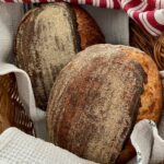 TWO LOAVES OF SAME DAY SOURDOUGH BREAD NESTLED IN A CANE BASKET WITH A RED AND WHITE DISH TOWEL