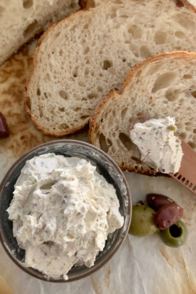 Olive cream cheese spread served in a small bowl. There is a rose gold butter knife and sourdough bread in the photo.