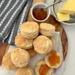 Buttery and flaky sourdough biscuits displayed on a wooden paddle smothered in apricot jam and butter.