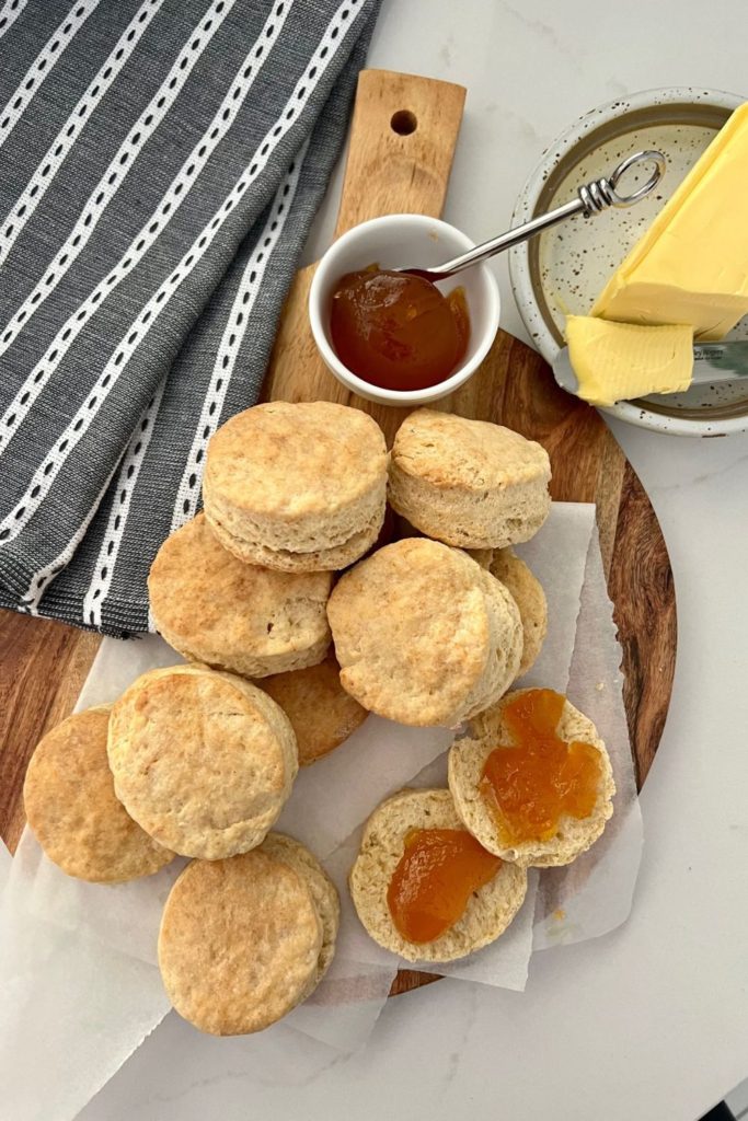 Flaky sourdough biscuits served on a wooden paddle. There is a striped dish towel on the left and a small dish of apricot jam and butter.