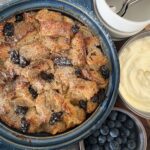 Sourdough bread pudding with raisins baked in a blue casserole dish. It is on a wooden tray with a dish of whipped cream and a bowl of blueberries.