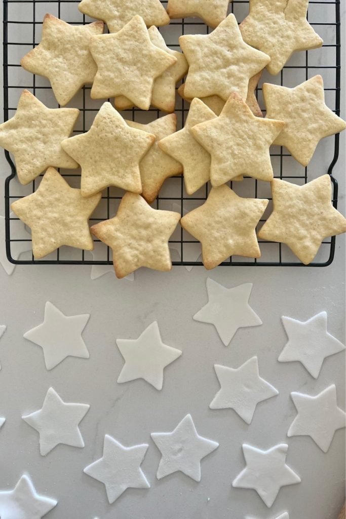 Baked sourdough sugar cookies in the shape of a star sitting on a black wire rack. There are star shaped pieces of fondant underneath waiting to be stuck to the cookies.