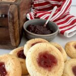 Sourdough thumbprint cookies sitting in front of a bowl of raspberry jam. There is a red and white dish cloth in the background and a wooden box.