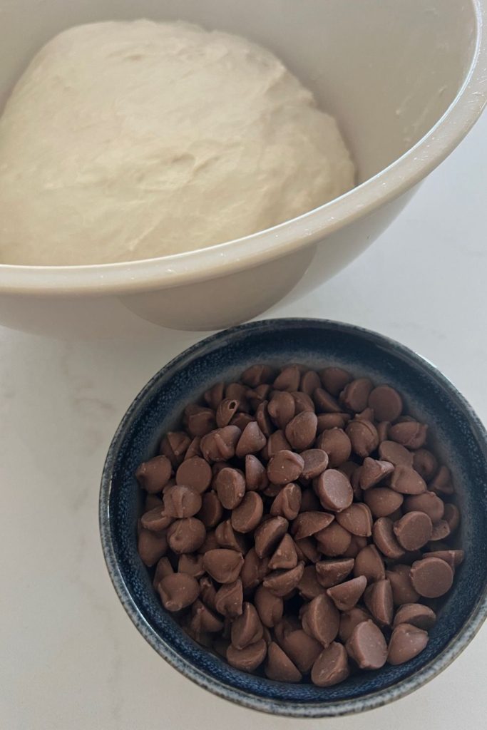 A bowl of sourdough bread dough sitting next to a blue rimmed bowl of chocolate chips.