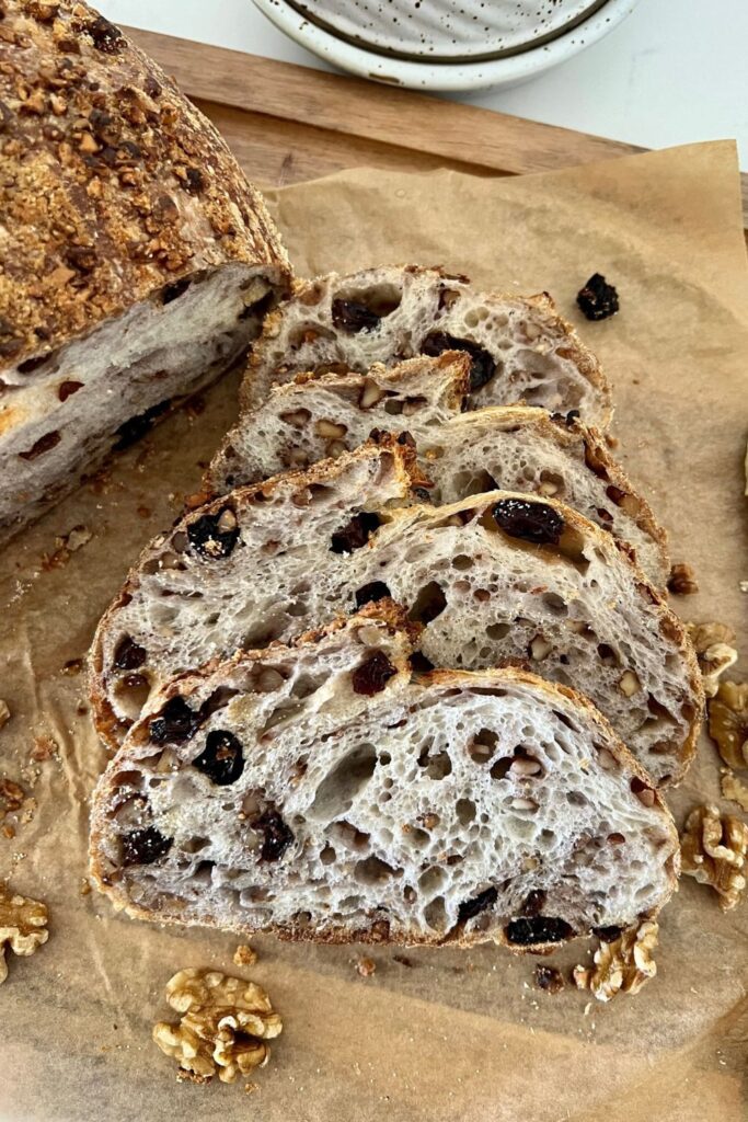 A loaf of walnut raisin sourdough bread that has been sliced and is sitting on a piece of parchment paper.