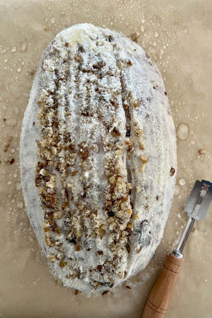 Walnut Raisin Sourdough that has been flipped out of the banneton onto a piece of parchment. The dough has been scored down the right side and there is a lame sitting beside the dough which is crusted with walnuts.