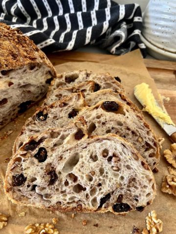 Walnut Raisin Sourdough Bread that has been sliced and arranged so you can see the "bunny" in the bread. There is a black and white dish towel sitting in the background and a wooden butter knife to the right of the sliced sourdough bread.