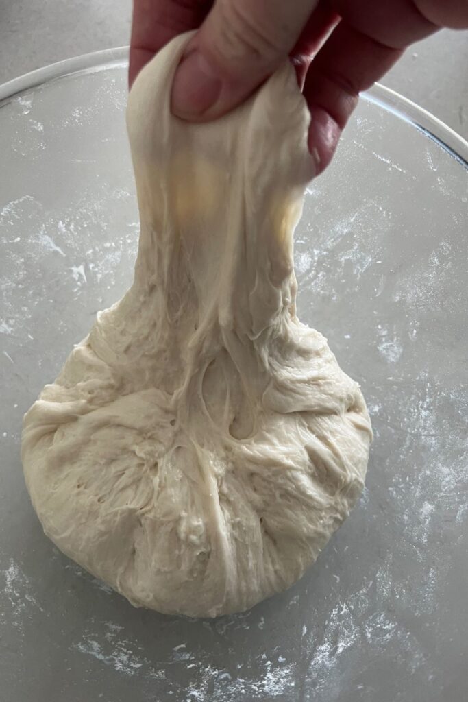 Sourdough bread dough sitting in a bowl with a hand stretching part of the dough upwards in a stretch and fold gluten development technique.