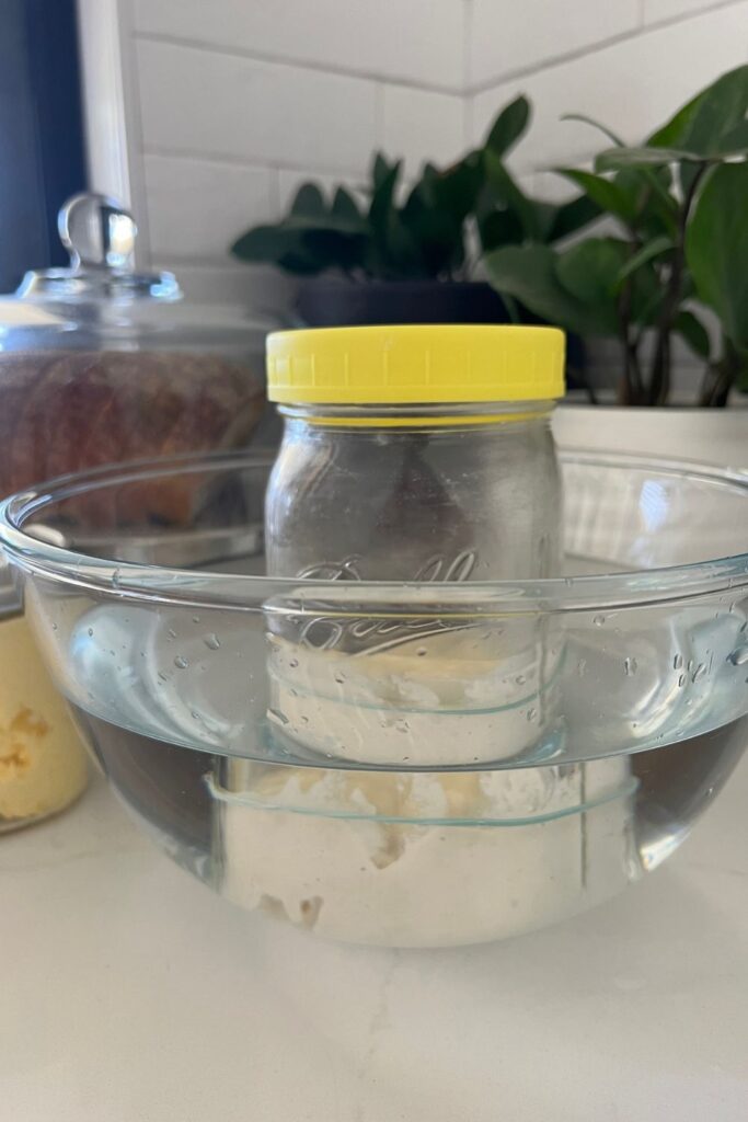 Jar of sourdough starter with a yellow lid sitting in a bowl of warm water. There are green plants behind the bowl and a loaf of sourdough bread and a jar of butter in the background.