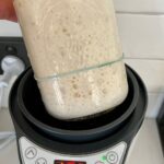 Jar of bubbly sourdough starter with a yellow lid being placed in an electric yogurt maker. It says 10hrs on the LED screen.