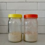Two jars of sourdough starter sitting on a white granite bench in front of a white tiled wall. The jar on the left has a yellow lid the sides of the jar are clean. The jar on the right has an orange lid and the sides of the jar are dirty.