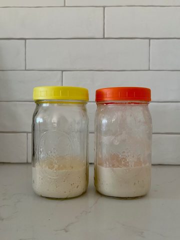 Two jars of sourdough starter sitting on a white granite bench in front of a white tiled wall. The jar on the left has a yellow lid the sides of the jar are clean. The jar on the right has an orange lid and the sides of the jar are dirty.
