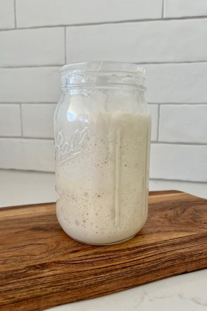 Tall mason jar of sourdough starter sitting on a wooden board in front of a white tiled wall. The sourdough starter has been fed and is rising inside the jar.