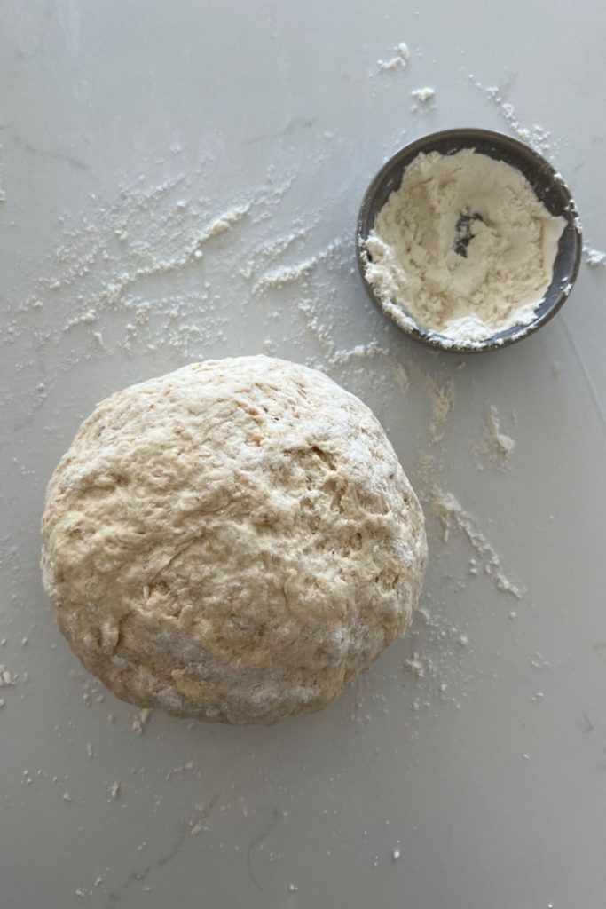 Sourdough Soda Bread dough formed in a rough ball. There is flour on the counter top and a small bowl of flour sitting to the right of the dough.