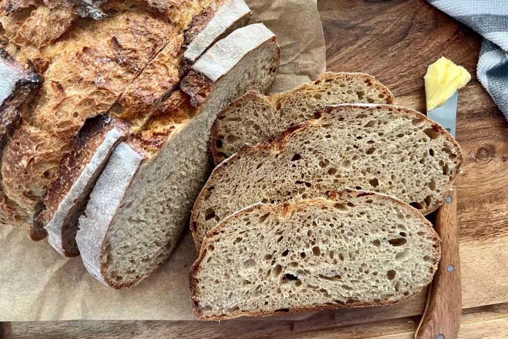Sourdough rye bread that has been sliced across the boule. The slices are laid out on a wooden board to reveal the soft crumb. There is a butter knife on the side of the slices of sourdough.