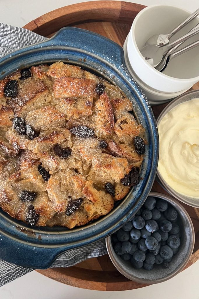 Bread Pudding using leftover sourdough bread - served in a blue oven dish surrounded by blueberries and whipped cream.