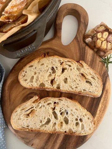 Roasted Garlic Sourdough Bread that has been sliced. There are 2 slices sitting on a round wooden board, as well as a head of roasted garlic in the background.