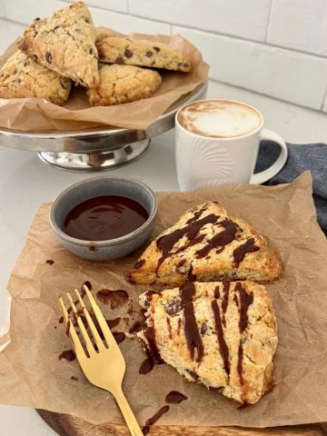 Two sourdough chocolate chip scones drizzled with chocolate ganache. There is a plate of scones in the background, as well as a coffee in a white mug.