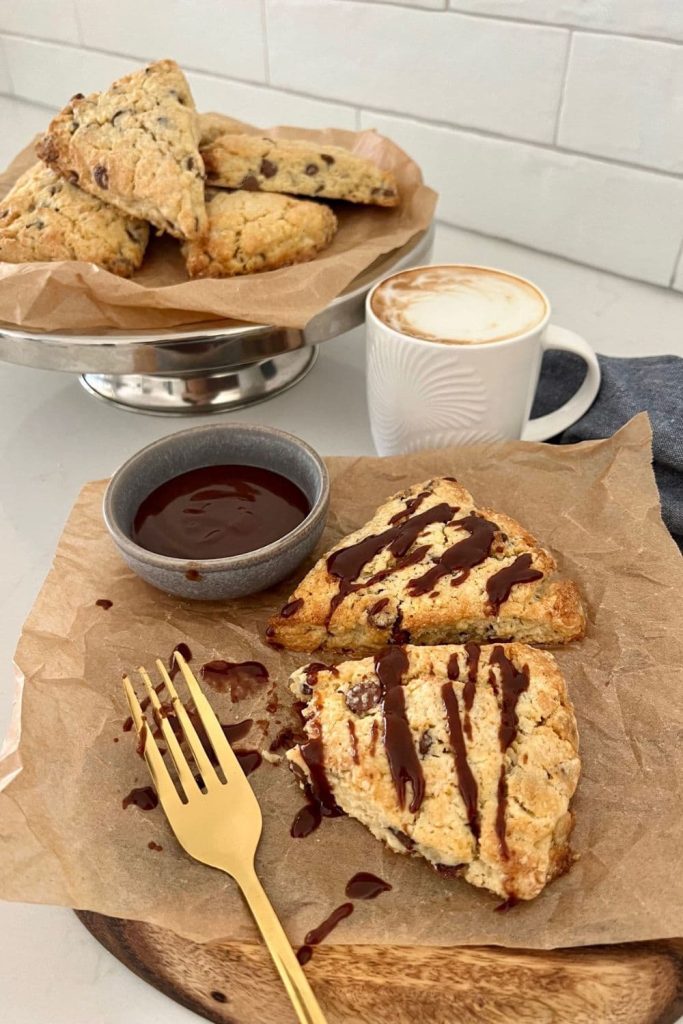 Two sourdough chocolate chip scones drizzled with chocolate ganache. There is a plate of scones in the background, as well as a coffee in a white mug.