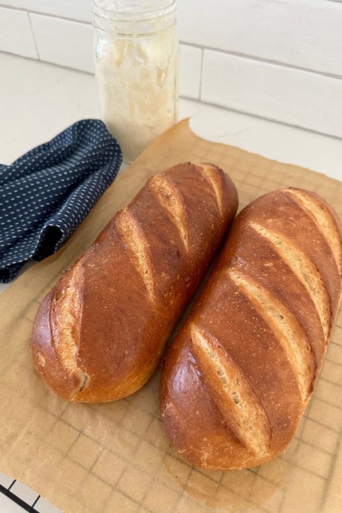 Two loaves sourdough French Bread sitting on a piece of parchment paper on top of a wire cooling rack. There is a navy blue dish towel and jar of sourdough starter in the background.