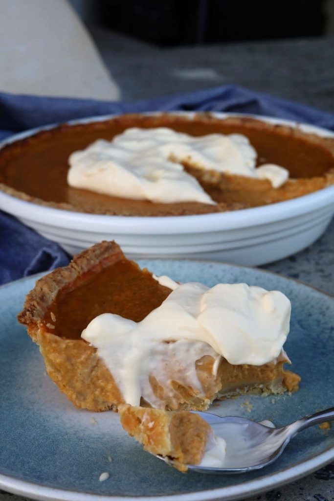 A slice of sourdough pumpkin pie served on a blue stoneware plate topped with whipped cream. You can see the rest of the sourdough pumpkin pie in the background of the photo.